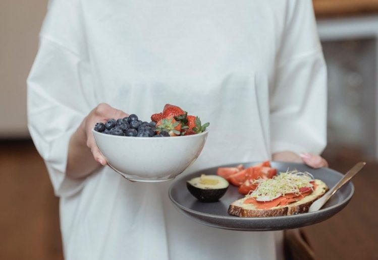 Person holding a bowl of fresh berries and a plate with avocado toast, displaying a healthy breakfast spread.