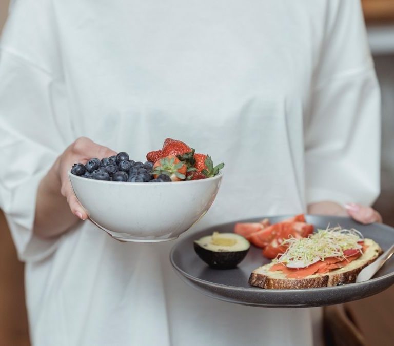 Person holding a bowl of fresh berries and a plate with avocado toast, displaying a healthy breakfast spread.