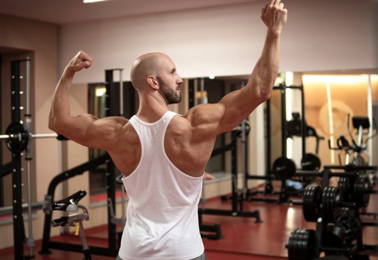 A muscular man in a gym flexes his back muscles wearing a white tank top.
