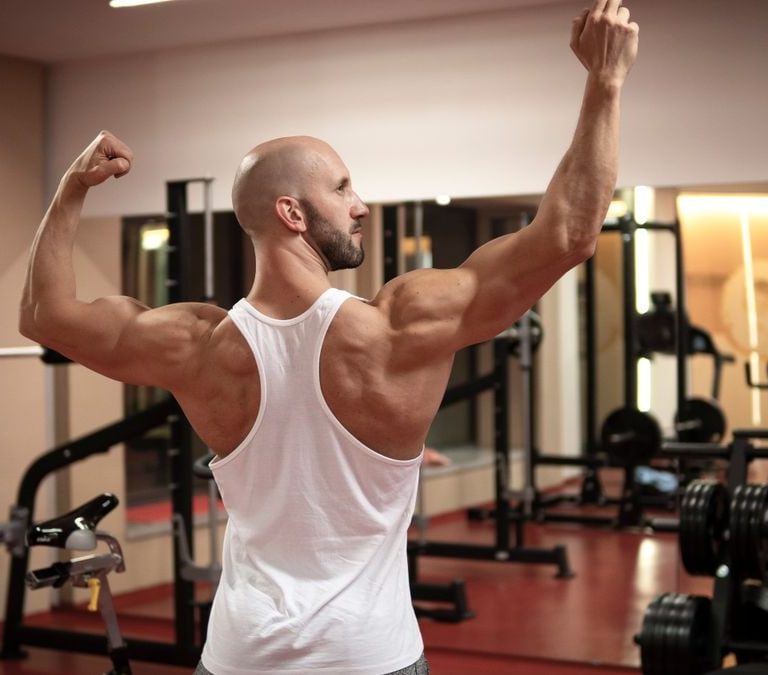 A muscular man in a gym flexes his back muscles wearing a white tank top.